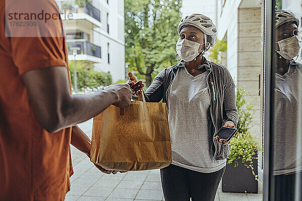 Frau mit schützender Gesichtsmaske übergibt einem Mann an der Tür eine Lebensmitteltüte