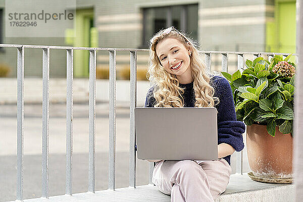 Glückliche junge blonde Frau mit Laptop sitzt auf dem Wandbuch