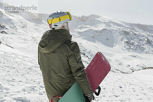 Junger Mann mit Snowboard steht auf schneebedecktem Berg