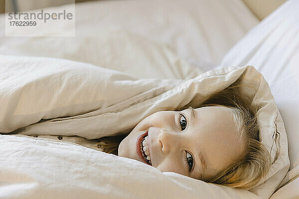 Smiling girl wrapped in blanket lying on bed at home