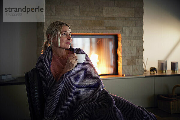 Blonde Frau  in eine Decke gehüllt  hält eine Kaffeetasse in der Hand und sitzt zu Hause