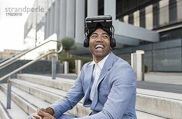 Fröhlicher Geschäftsmann mit Virtual-Reality-Simulator sitzt vor dem Gebäude