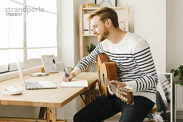 Lächelnder junger Mann mit Gitarre  der zu Hause Musiknoten schreibt und am Laptop lernt