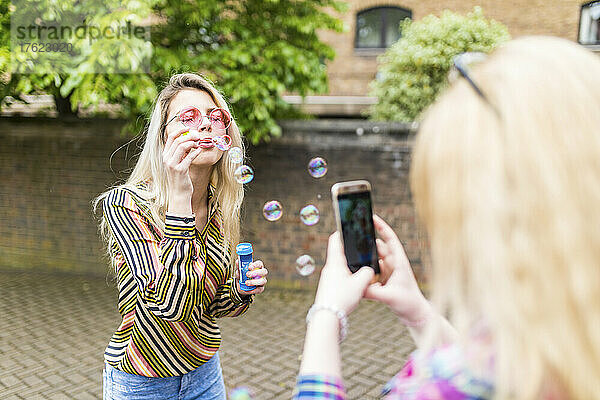 Teenager photographing friend blowing soap bubbles through smart phone