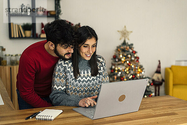 Mann umarmt lächelnde Freundin  die zu Hause Laptop benutzt