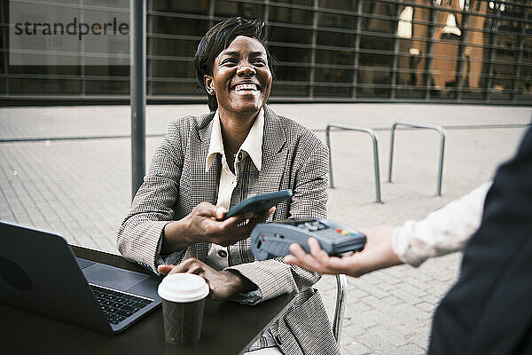 Glückliche Geschäftsfrau  die kontaktlos per Smartphone an die Kellnerin im Straßencafé bezahlt