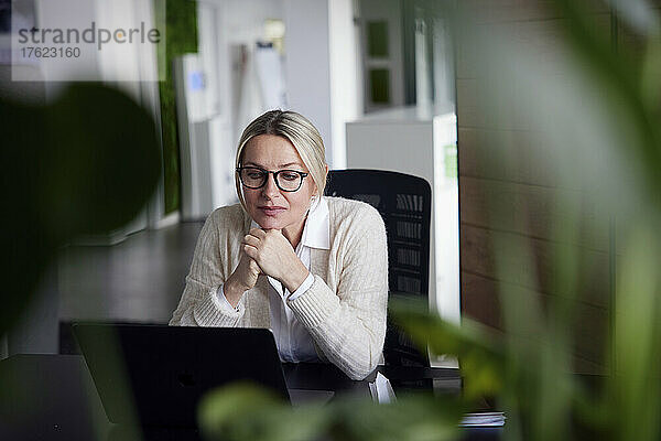 Nachdenkliche Geschäftsfrau mit den Händen am Kinn und Blick auf den Laptop im Büro