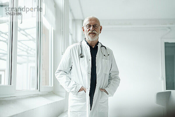 Doctor standing with hands in pockets by window at hospital