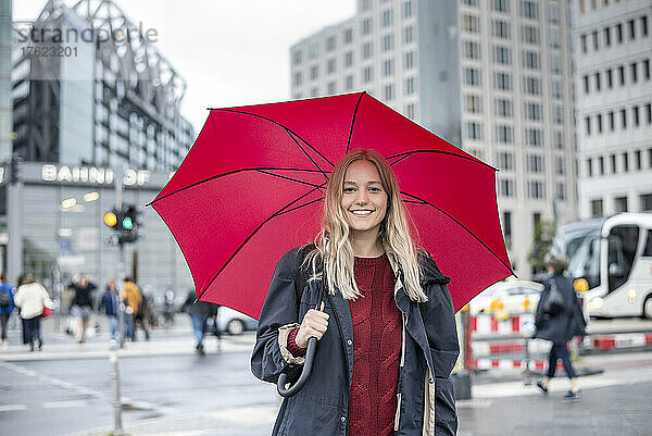 Lächelnde junge Frau steht mit rotem Regenschirm auf der Straße