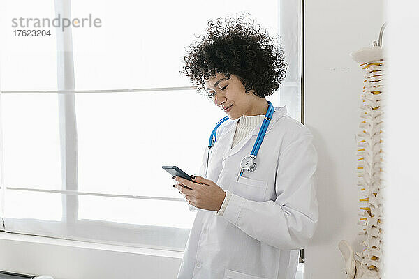 Doctor text messaging through mobile phone standing by window in medical office