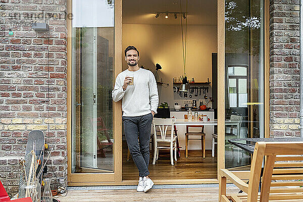 Lächelnder Mann mit einem Glas Kaffee lehnt an der Tür