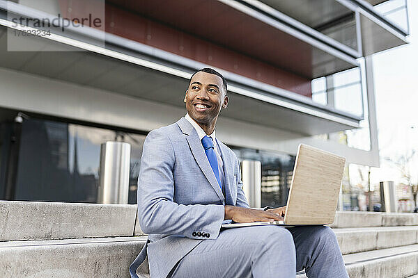 Glücklicher Geschäftsmann mit Laptop  der auf Stufen vor dem Gebäude sitzt