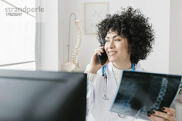 Lächelnder Arzt mit Röntgenbild  der auf dem Smartphone spricht und in der medizinischen Klinik sitzt