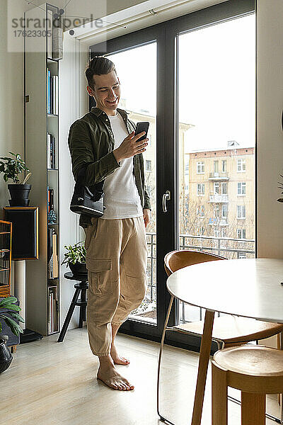 Glücklicher junger Mann  der zu Hause sein Smartphone am Fenster benutzt