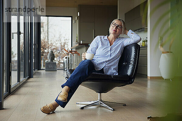 Entspannte Frau mit Brille und Kaffeetasse sitzt zu Hause auf einem Stuhl