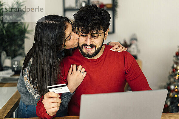 Junge Frau küsst ihren Freund auf die Wange  der zu Hause mit dem Laptop online einkauft
