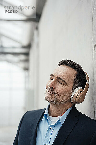 Geschäftsmann mit geschlossenen Augen hört Musik über kabellose Kopfhörer an der Wand