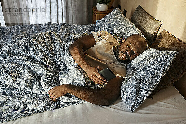 Man using smart phone lying on bed in morning at home