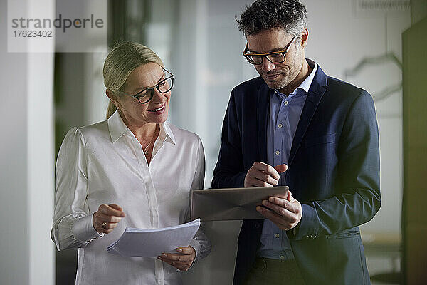 Lächelnde Geschäftsfrau hält ein Dokument in der Hand und diskutiert mit einem Kollegen über einen Tablet-PC im Büro