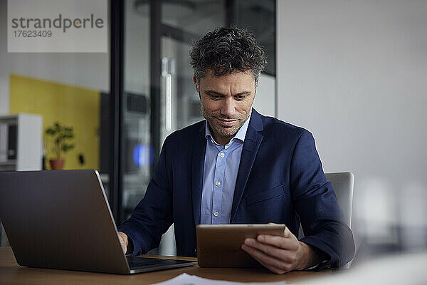 Lächelnder Geschäftsmann mit Tablet-PC und Laptop am Schreibtisch im Büro