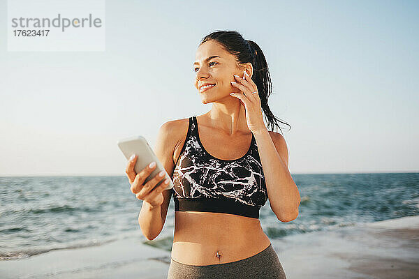 Lächelnde Frau hört am Strand Musik über kabellose In-Ear-Kopfhörer