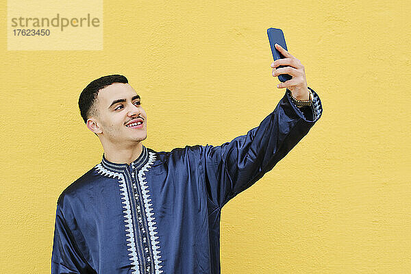 Smiling young man photographing through mobile phone in front of wall