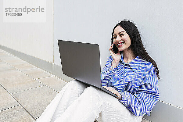 Lächelnde Geschäftsfrau mit Laptop  die vor der Wand telefoniert