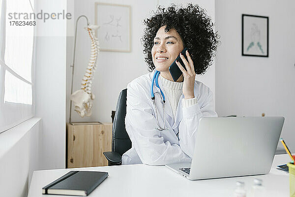 Lächelnder junger Arzt sitzt mit Laptop und telefoniert in der Klinik