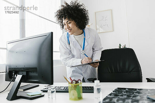 Lächelnder Arzt mit Klemmbrett und Blick auf den Desktop-PC in der Klinik