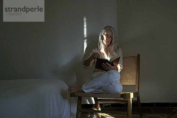 Frau liest ein Buch und sitzt zu Hause auf einem Stuhl neben dem Bett