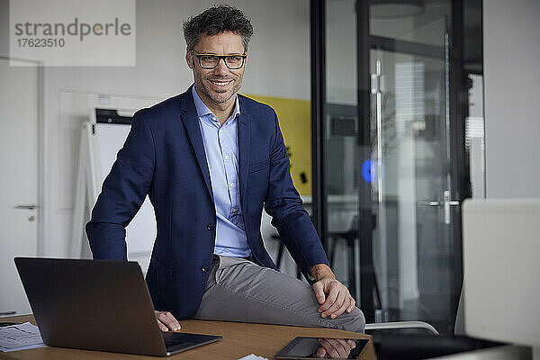 Glücklicher Geschäftsmann mit Brille sitzt mit Laptop auf dem Schreibtisch im Büro