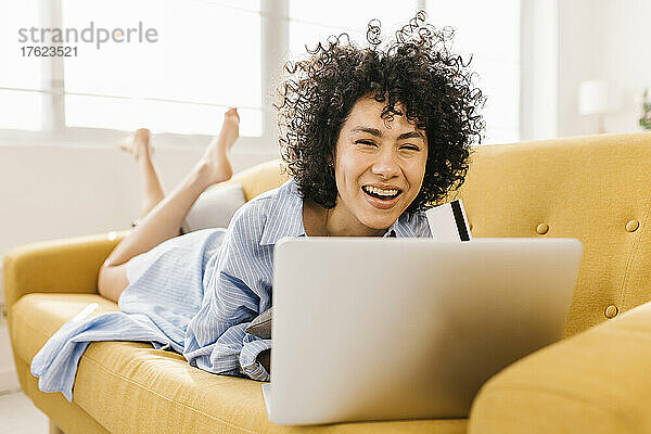 Fröhliche junge Frau mit Kreditkarte und Laptop liegt zu Hause auf dem Sofa im Wohnzimmer
