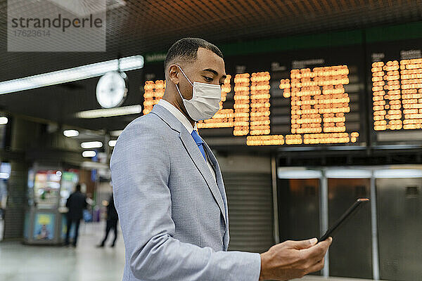 Geschäftsmann mit Schutzmaske und Tablet-PC neben der Abfahrtstafel am Bahnhof
