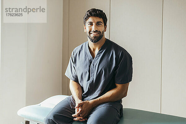Lächelnder junger Physiotherapeut sitzt auf einer Trage in der medizinischen Klinik