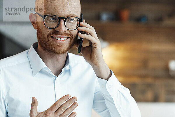 Lächelnder junger Geschäftsmann mit Brille  der im Büro auf dem Smartphone spricht
