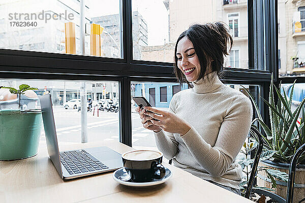 Glückliche Geschäftsfrau mit Laptop und Smartphone sitzt im Café