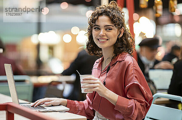 Lächelnder Freiberufler mit Einweg-Kaffeetasse und Laptop im Café