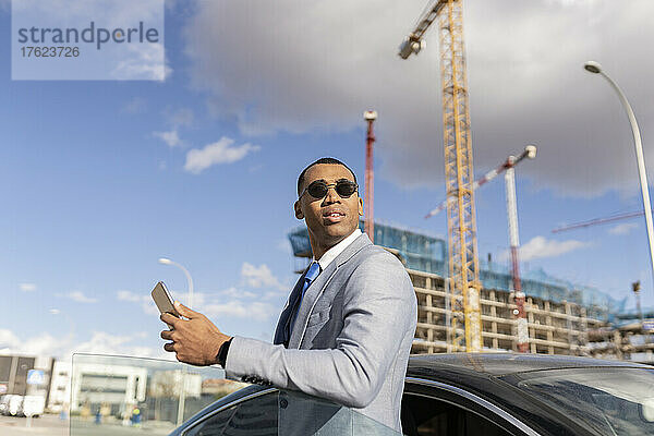Geschäftsmann mit Sonnenbrille und Smartphone  der an einem sonnigen Tag am Auto steht