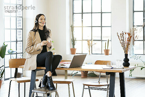 Lächelnde Geschäftsfrau  die über ein Headset spricht und am Tisch im Heimbüro sitzt