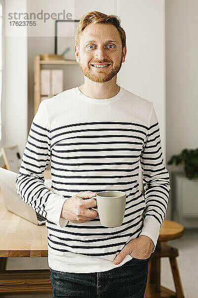 Lächelnder Mann steht mit der Hand in der Tasche und hält eine Kaffeetasse zu Hause