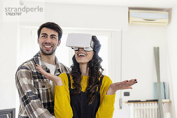 Smiling woman with VR glasses gesturing by boyfriend at new home