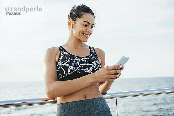 Happy young woman surfing net through mobile phone standing by railing