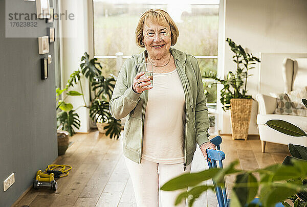 Glückliche ältere Frau mit einem Glas Wasser  die zu Hause neben dem Stuhl steht
