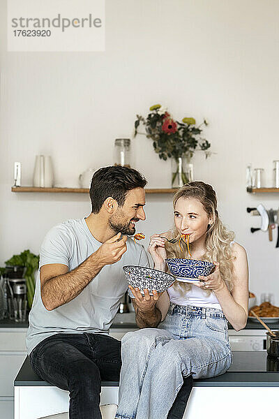 Lächelnder junger Mann schaut seiner Freundin zu  die zu Hause auf der Kücheninsel Spaghetti isst