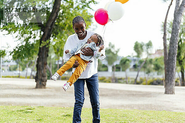 Verspielte Mutter trägt Tochter mit Luftballons im Park