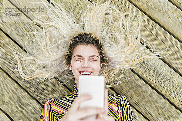 Cheerful teenager taking selfie through mobile phone lying on floorboard