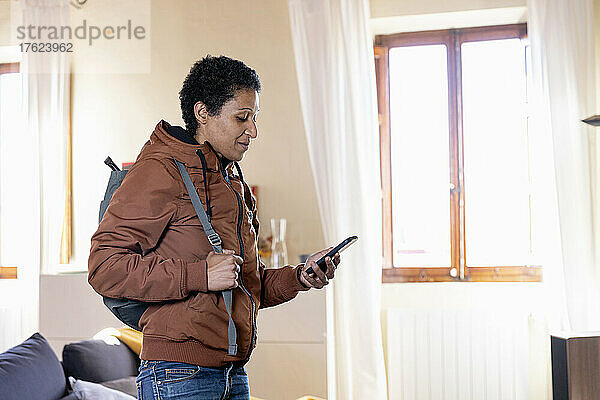 Frau mit Rucksack benutzt Mobiltelefon im heimischen Wohnzimmer