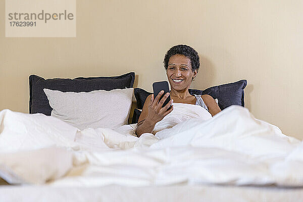 Lächelnde Frau  die zu Hause im Bett ihr Smartphone überprüft