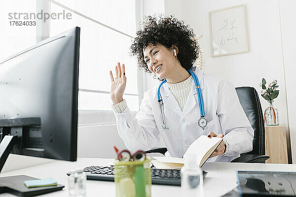 Lächelnder Arzt winkt bei Videoanruf über Desktop-PC im Krankenhaus mit der Hand