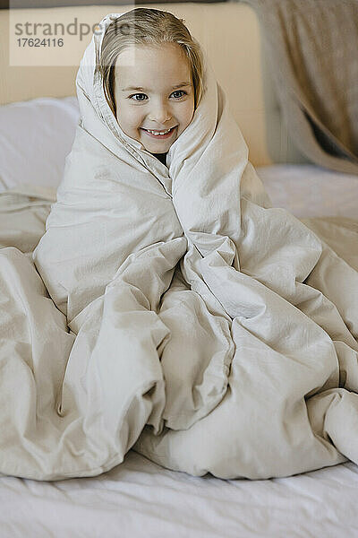 Lächelndes süßes blondes Mädchen  in eine Decke gehüllt  sitzt zu Hause auf dem Bett
